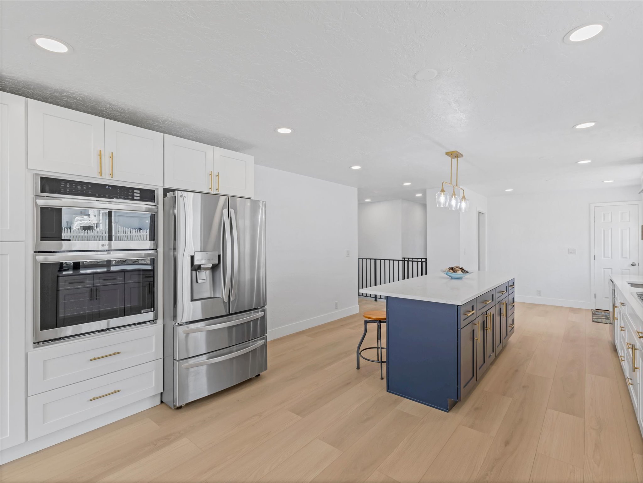 Modern kitchen featuring stainless steel appliances, white cabinetry with gold-tone hardware, a blue kitchen island with a light-toned countertop, recessed lighting, and wood-finish flooring