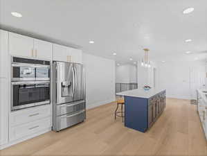 Modern kitchen featuring stainless steel appliances, white cabinetry with gold-tone hardware, a blue kitchen island with a light-toned countertop, recessed lighting, and wood-finish flooring