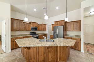 Kitchen featuring wood finish cabinetry, stainless steel appliances, light stone countertops, an island with sink, and pendant lighting
