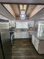 Kitchen featuring a wooden tray ceiling, white cabinetry, stainless steel appliances, light stone counters, and dark wood-style floors