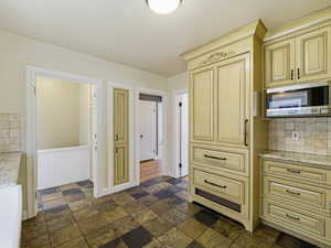 Kitchen with cream cabinetry, light stone countertops, stainless steel microwave, stone tile flooring, and wainscoting