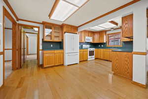 Kitchen with white appliances, wood finish cabinetry, crown molding, dark countertops, and light wood finished floors
