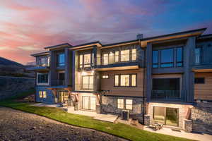 Back of property at dusk featuring a balcony, stone siding, and a lawn