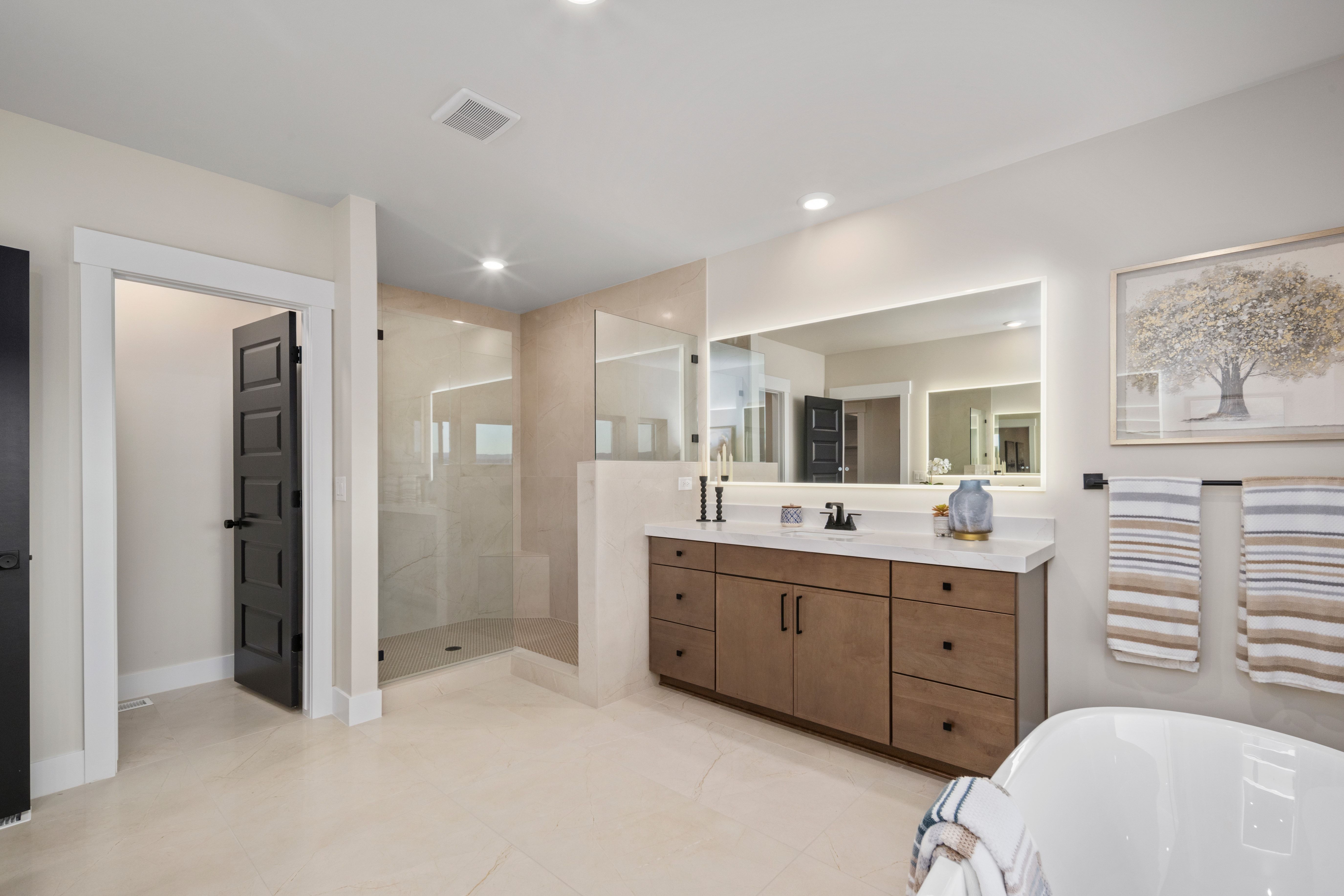 Spacious bathroom featuring a dual vanity with wood-finish cabinetry, a large illuminated mirror, a glass-enclosed shower, and light-toned tile flooring
