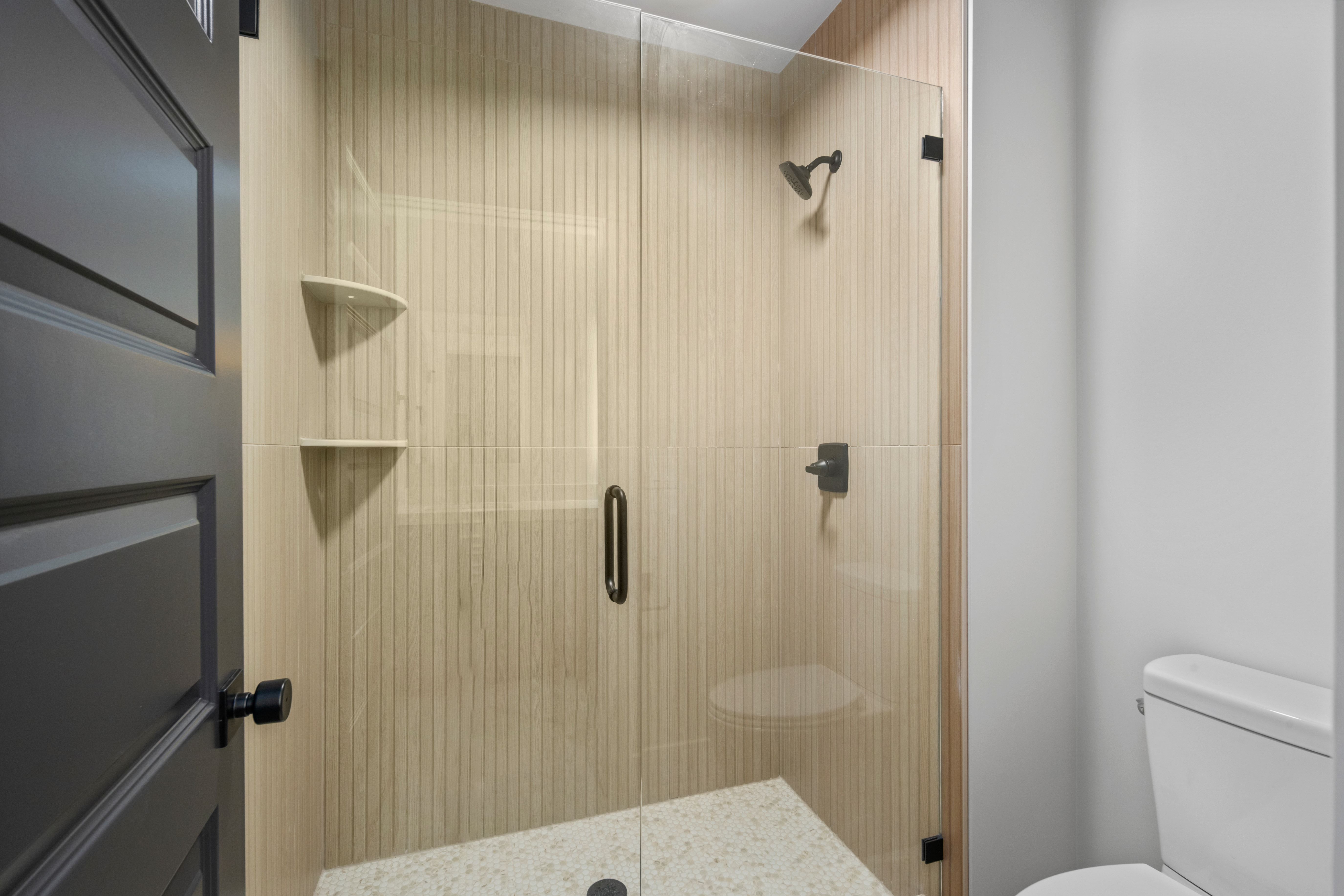 Glass-enclosed shower featuring vertically textured wall tiles, matte black fixtures, and integrated corner shelves