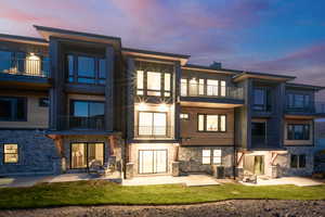 Back of house at dusk featuring a balcony, a patio area, stone siding, and a yard