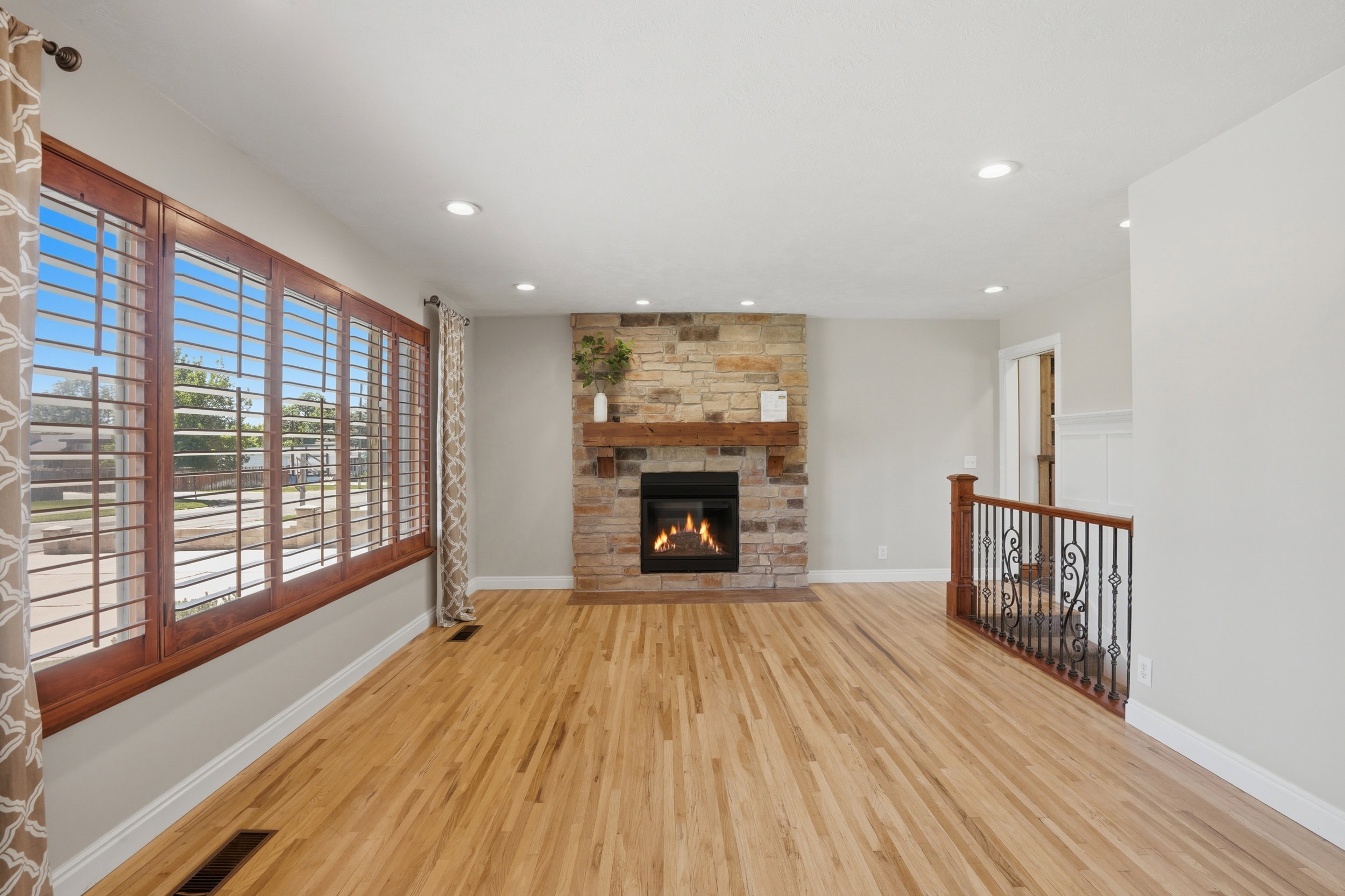 Living area featuring wood-finish flooring, a stone-clad fireplace with a wood mantel, and expansive windows with plantation shutters