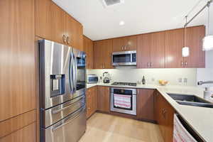 Kitchen with stainless steel appliances, wood finish cabinets, decorative light fixtures, light stone counters, and light wood-style floors