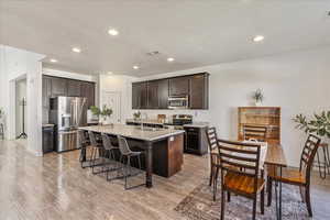 Kitchen featuring stainless steel appliances, dark wood finish cabinets, light stone countertops, a kitchen island with sink, and light wood-style flooring