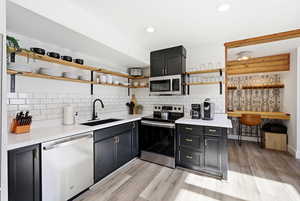 Kitchen featuring dark cabinets, stainless steel appliances, open shelves, decorative backsplash, and light wood finished floors