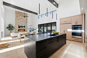 Kitchen featuring a large island with sink, a stone fireplace, light wood-type flooring, stainless steel double oven, and modern cabinets