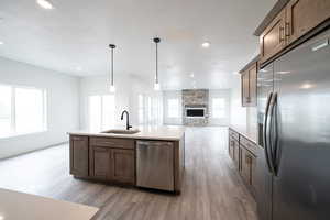 Kitchen featuring open floor plan, stainless steel appliances, light wood-style floors, a stone fireplace, and a kitchen island with sink