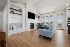 Living room featuring built in shelves, vaulted ceiling, a ceiling fan, light wood finished floors, and a glass covered fireplace