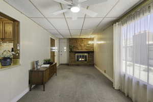 Living room featuring a drop ceiling, a stone fireplace, light carpet, wooden panel walls, and a ceiling fan