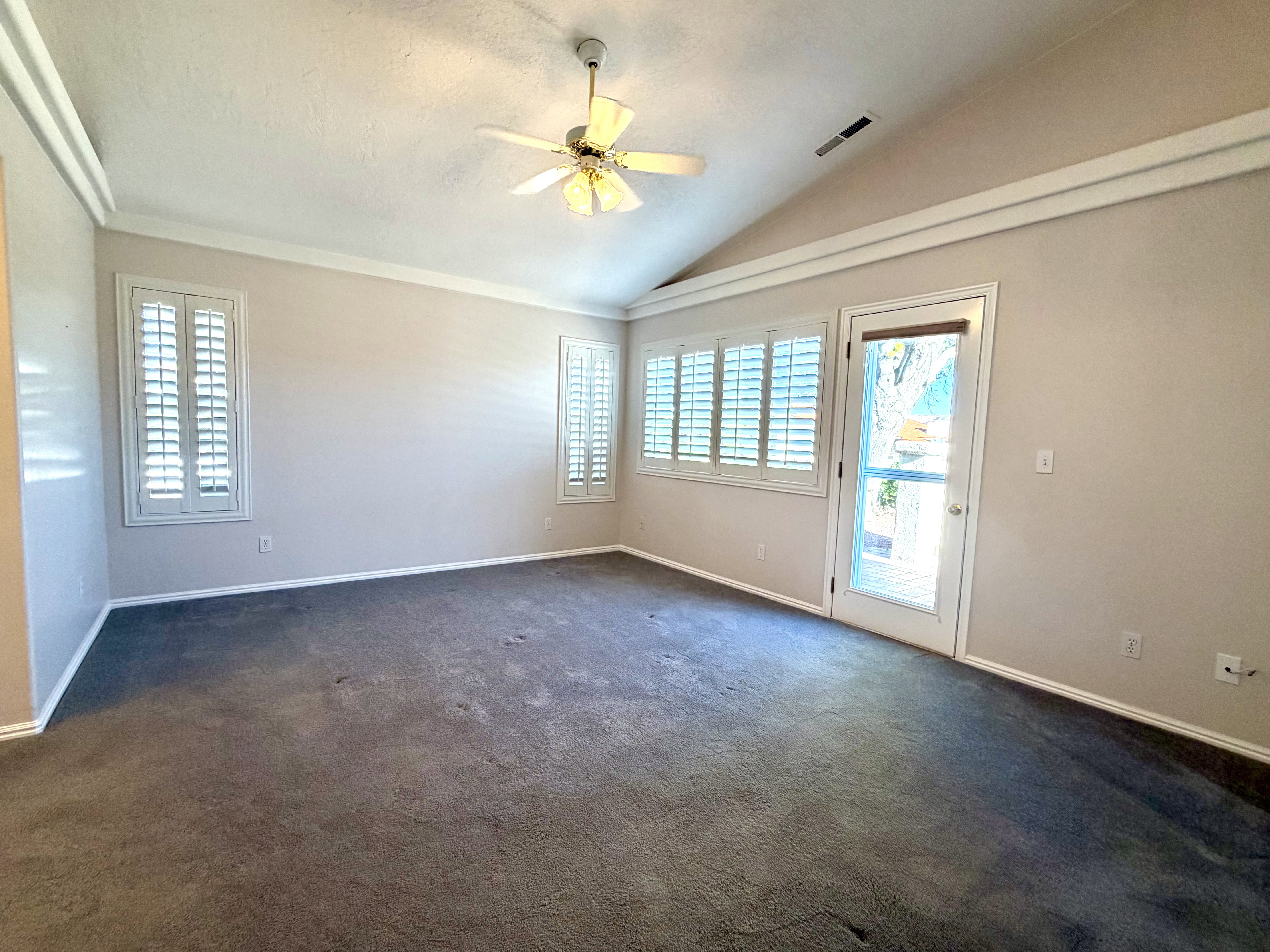 Spacious interior room featuring a vaulted ceiling with a ceiling fan, plush gray carpeting, white plantation shutters on multiple windows, and a glass-paneled door