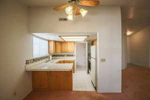 Kitchen with tile countertops, a peninsula, ceiling fan, freestanding refrigerator, and decorative backsplash