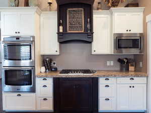 Kitchen with stainless steel appliances, white cabinetry, range hood, and light stone countertops