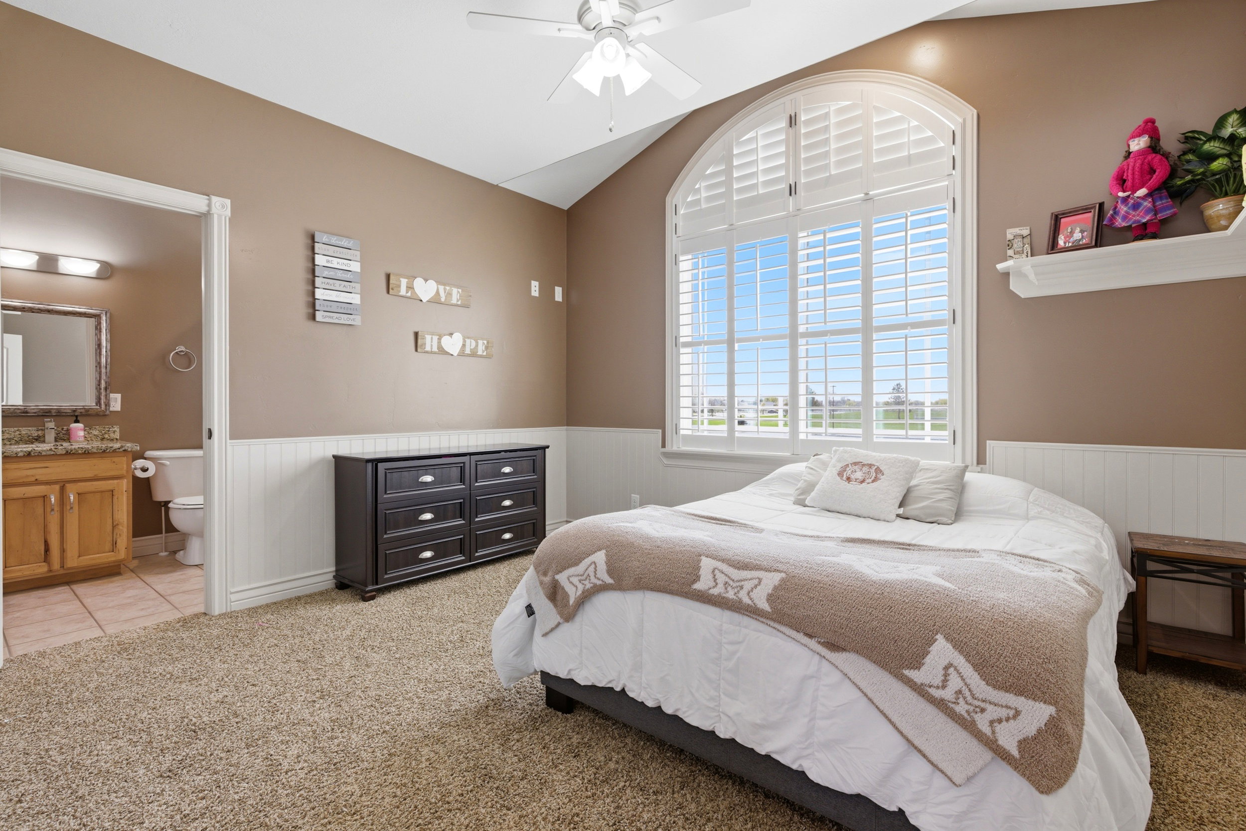 Bedroom featuring light colored carpet, lofted ceiling, a wainscoted wall, a ceiling fan, and ensuite bath