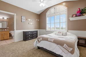 Bedroom featuring light colored carpet, lofted ceiling, a wainscoted wall, a ceiling fan, and ensuite bath