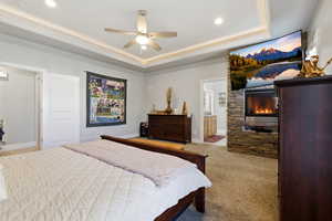Carpeted bedroom featuring a tray ceiling, a ceiling fan, and a glass covered fireplace