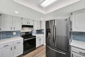 Kitchen with black range with electric cooktop, stainless steel fridge, tasteful backsplash, light wood-style floors, and white cabinetry