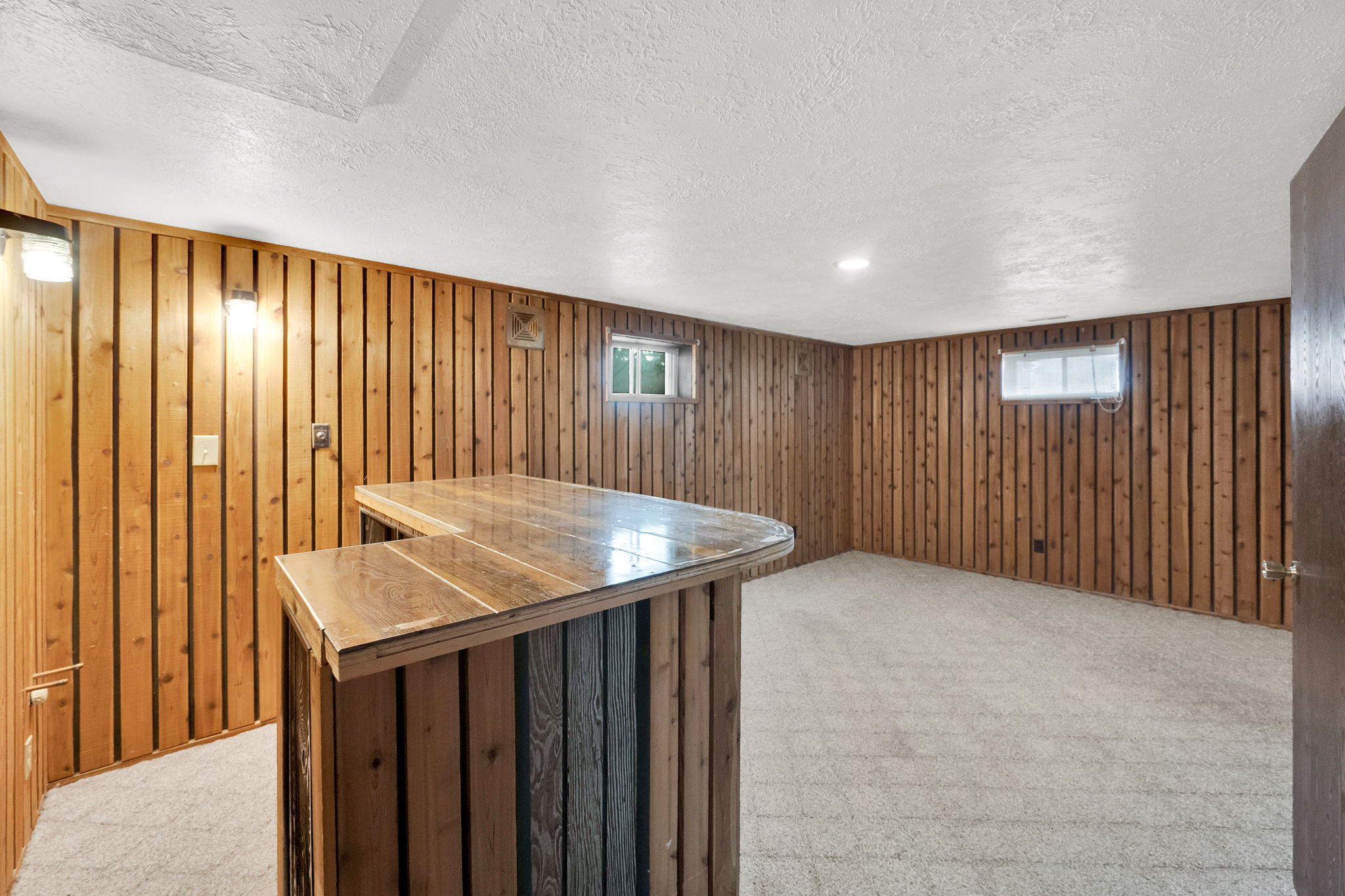 Basement with light colored carpet, a bar, plenty of natural light, wooden walls, and a textured ceiling