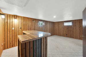 Basement with light colored carpet, a bar, plenty of natural light, wooden walls, and a textured ceiling