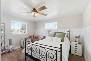 Bedroom featuring multiple windows, dark carpet, a ceiling fan, a decorative wall, and wainscoting