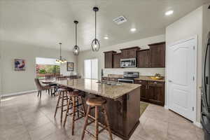 Kitchen with dark wood finish cabinetry, stainless steel appliances, light stone counters, a kitchen breakfast bar, and an island with sink