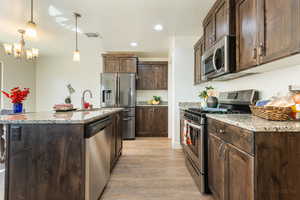Kitchen with stainless steel appliances, dark wood finish cabinets, light stone counters, a center island with sink, and light wood-style floors