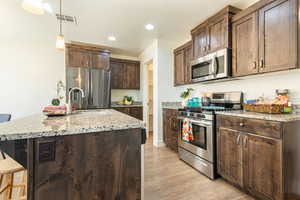 Kitchen with stainless steel appliances, dark wood finish cabinets, a center island with sink, light stone counters, and light wood-style flooring