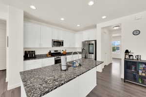 Kitchen featuring white cabinets, stainless steel appliances, a kitchen island with sink, dark stone counters, and dark wood-type flooring
