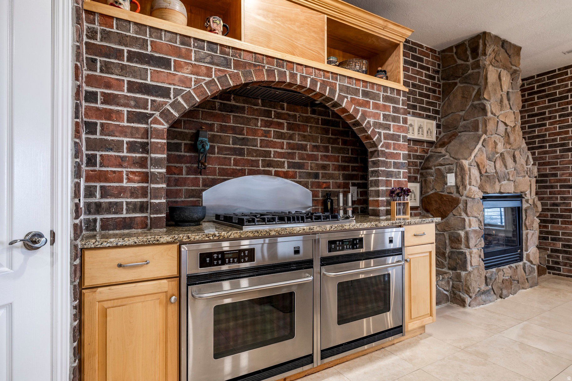 Kitchen with double oven, open shelves, dark stone countertops, a multi sided fireplace, and light wood finish cabinets