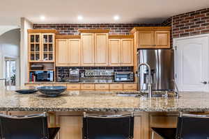 Kitchen featuring brick wall, stainless steel fridge, light stone counters, glass insert cabinets, and recessed lighting