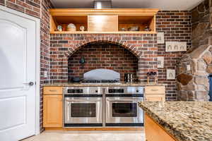 Kitchen featuring light stone counters, open shelves, double oven, brick wall, and light tile patterned flooring