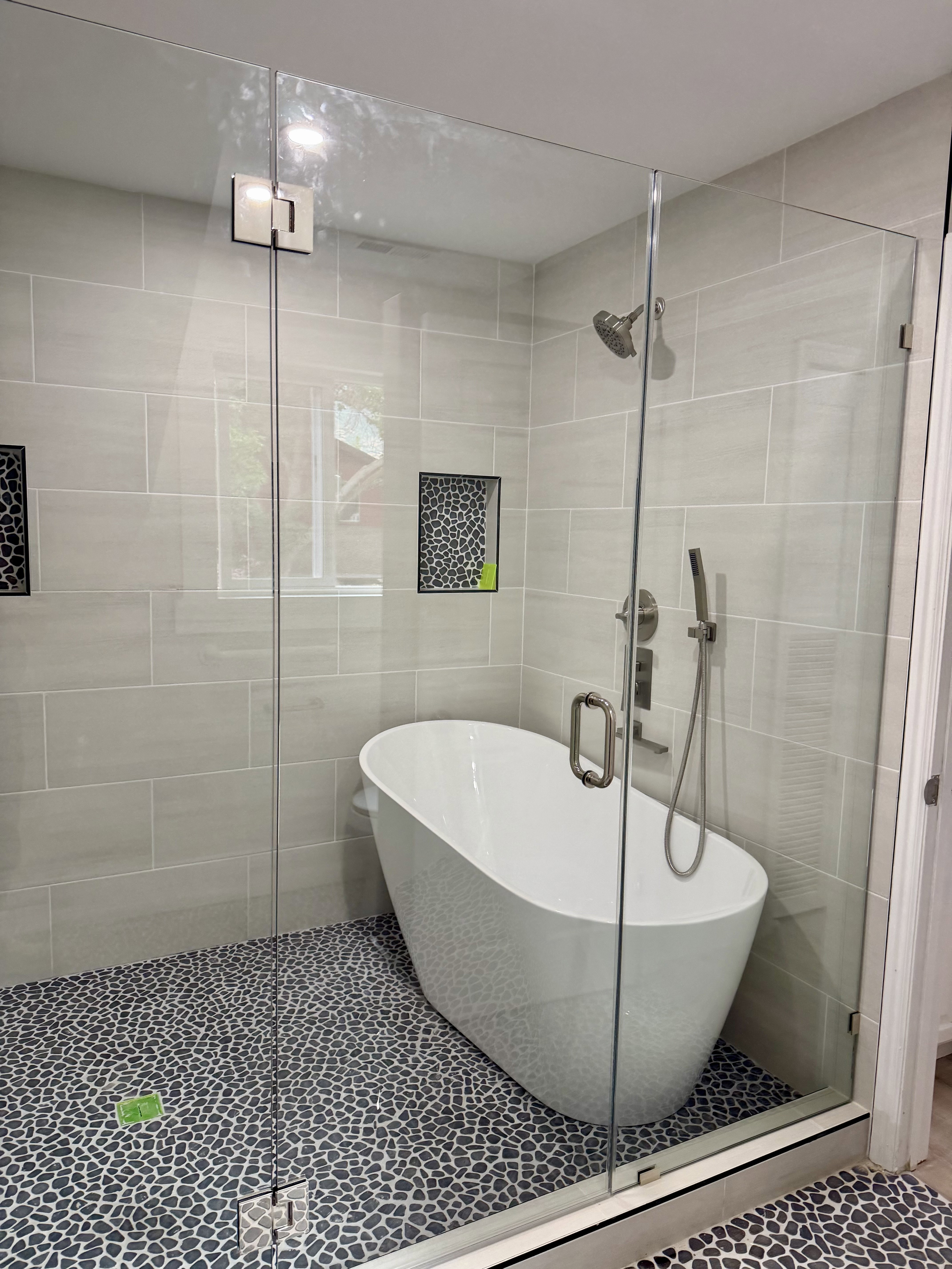 Glass-enclosed wet room featuring a freestanding soaking tub, pebble tile flooring, and light grey subway tile walls