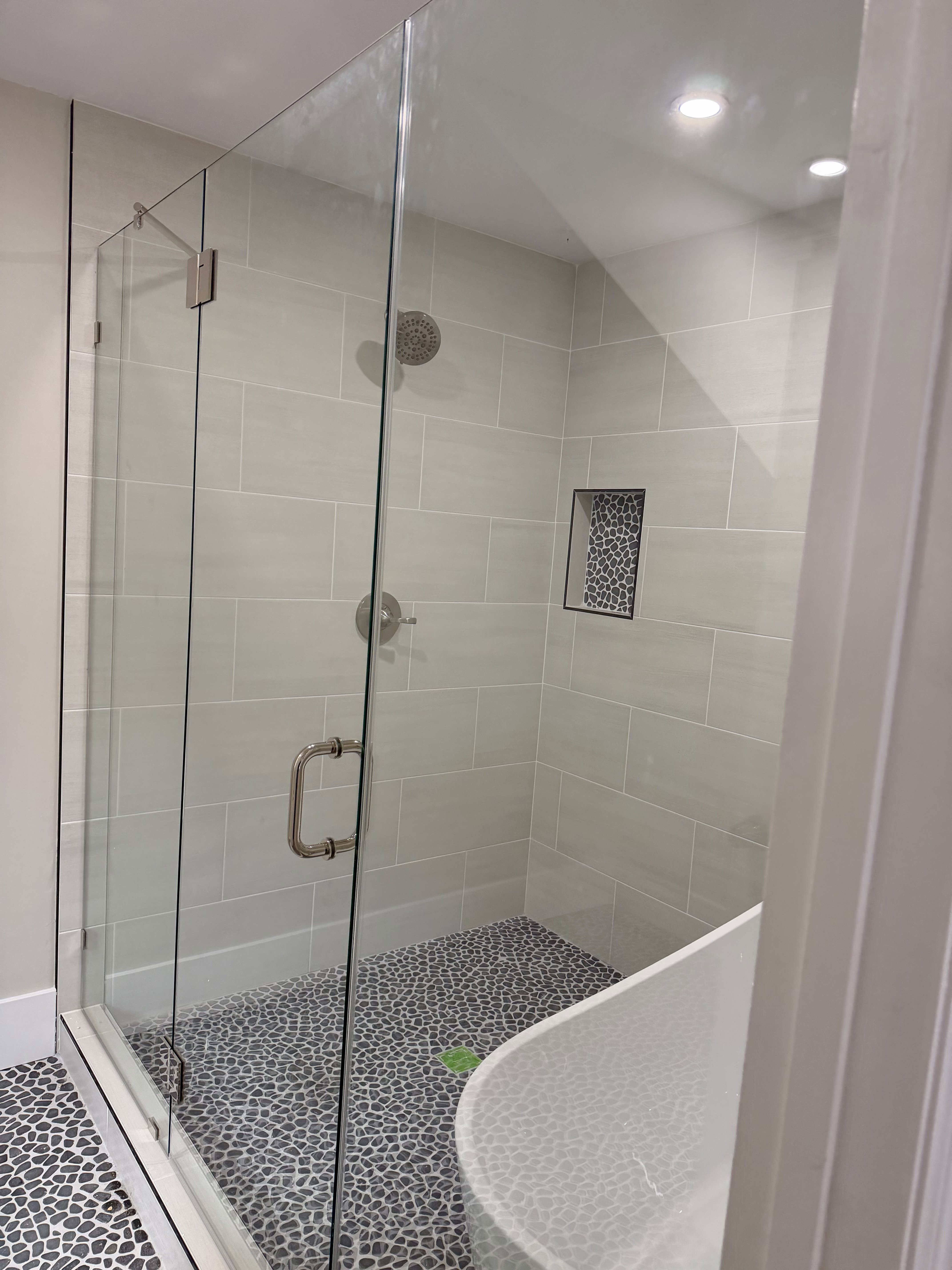 Walk-in shower with clear glass enclosure, light grey subway tile walls, a built-in shower niche with mosaic tile, and a pebble-finish floor
