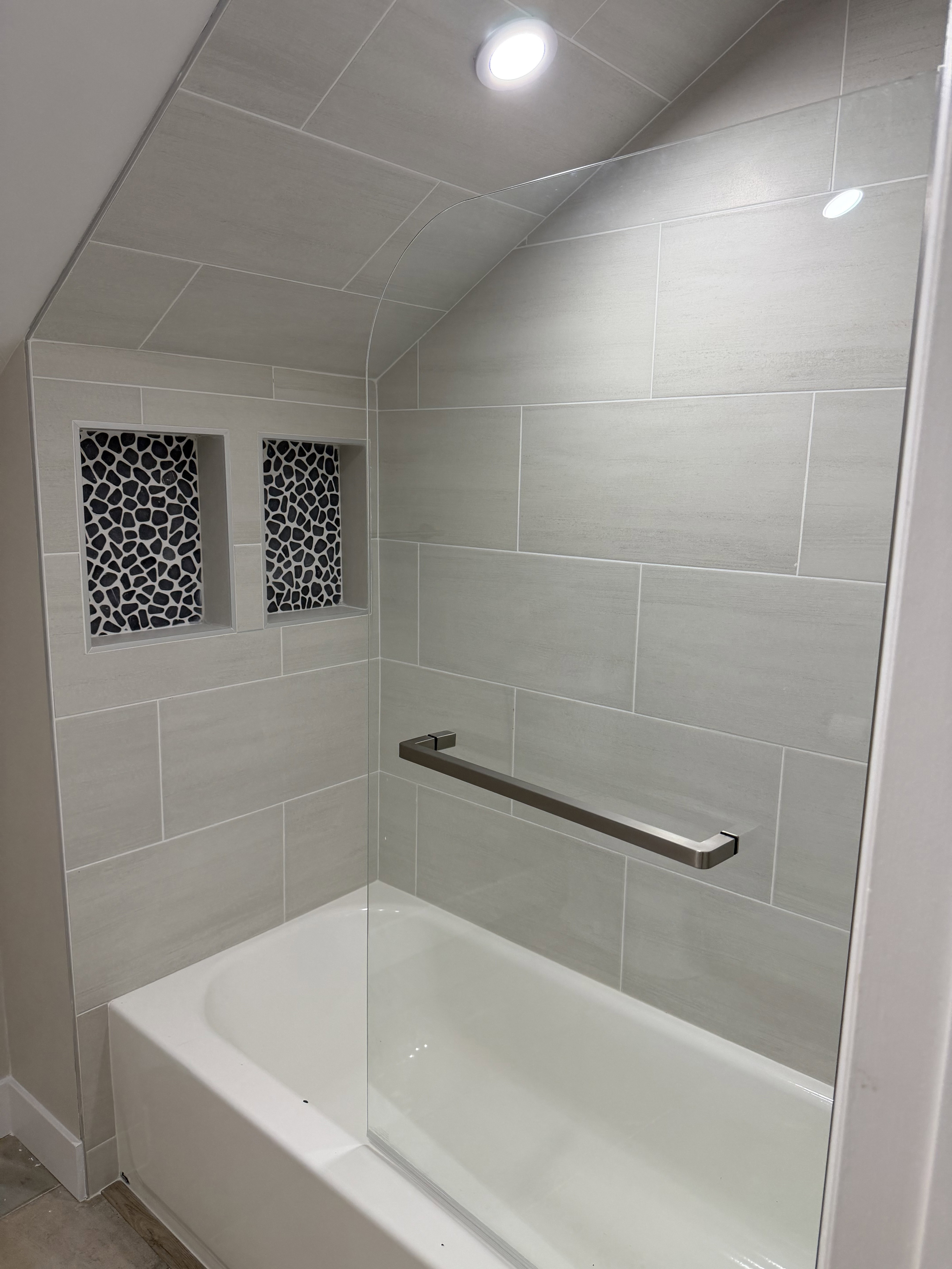 Bathtub with light gray subway tile surround, two tiled shower niches with black and white pebble mosaic, clear glass shower panel, and recessed lighting