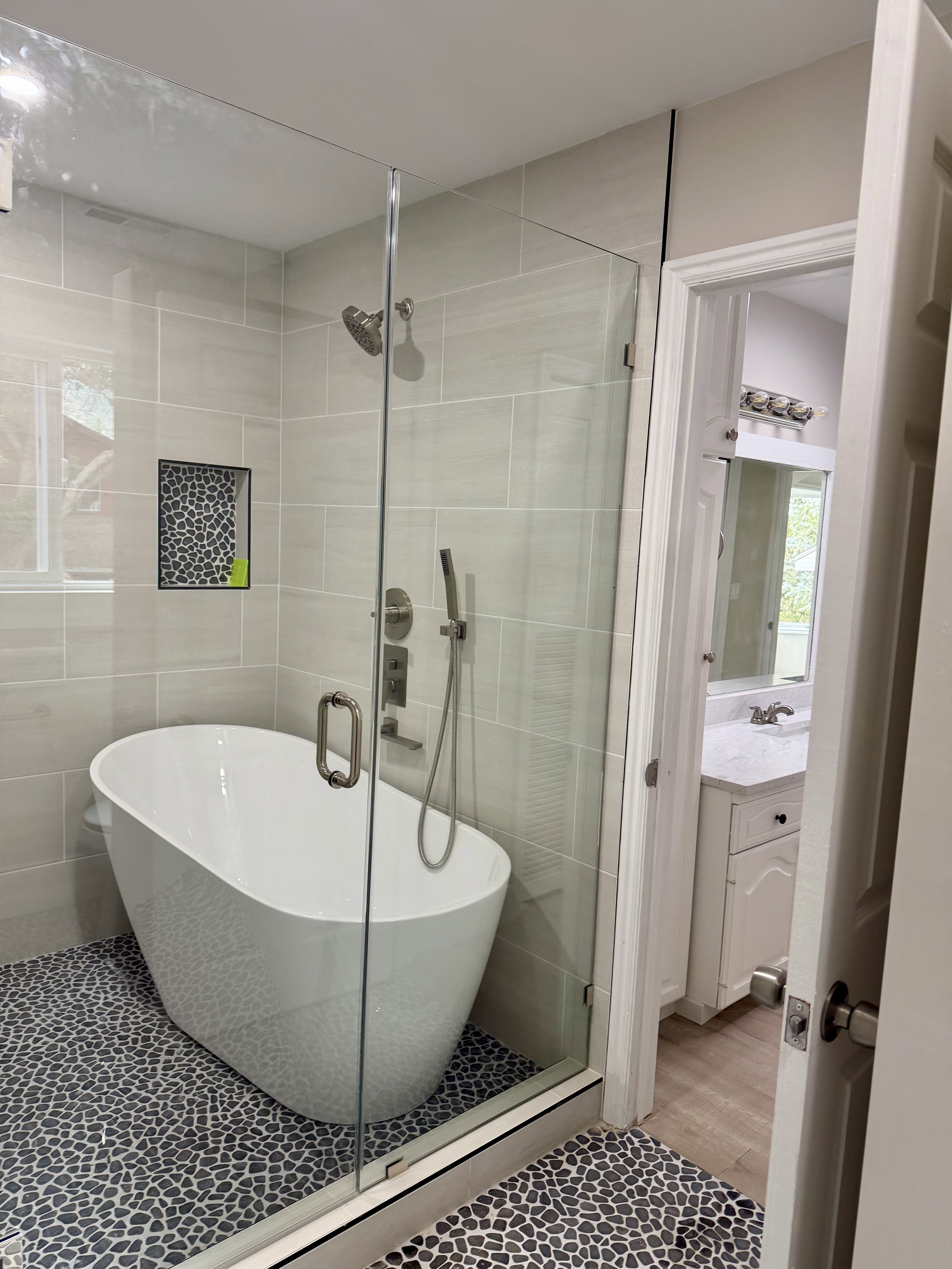 Enclosed shower featuring a freestanding soaking tub, pebble tile flooring, glass shower enclosure, a built-in shower niche, and a fixed showerhead with a handheld sprayer