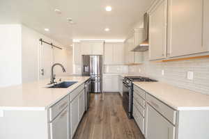 Kitchen featuring a barn door, stainless steel appliances, a center island with sink, dark wood-style flooring, and light stone countertops
