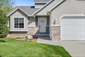 Doorway to property with brick siding, stucco siding, a lawn, concrete driveway, and a shingled roof