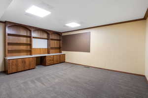 Unfurnished office featuring built in desk, dark colored carpet, wood walls, and ornamental molding