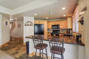 Kitchen featuring black appliances, a peninsula, light wood finish cabinetry, a kitchen bar, and dark stone finish flooring