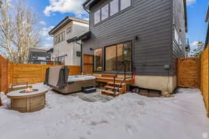 Snow covered back of property with a fenced backyard, a fire pit, and a hot tub