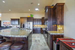Kitchen with a kitchen breakfast bar, an island with sink, stainless steel appliances, light stone countertops, and tasteful backsplash