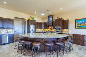 Kitchen with tasteful backsplash, a large island, light stone counters, stainless steel appliances, and dark wood finish cabinetry