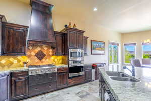 Kitchen with dark wood finish cabinetry, stainless steel appliances, light stone counters, backsplash, and a chandelier