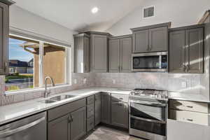 Kitchen with stainless steel appliances, gray cabinets, lofted ceiling, light stone counters, and backsplash