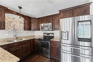 Kitchen featuring stainless steel appliances, dark wood finish cabinetry, hanging light fixtures, and light stone counters