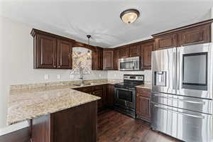 Kitchen with stainless steel appliances, dark wood finish cabinetry, a peninsula, light stone counters, and hanging light fixtures
